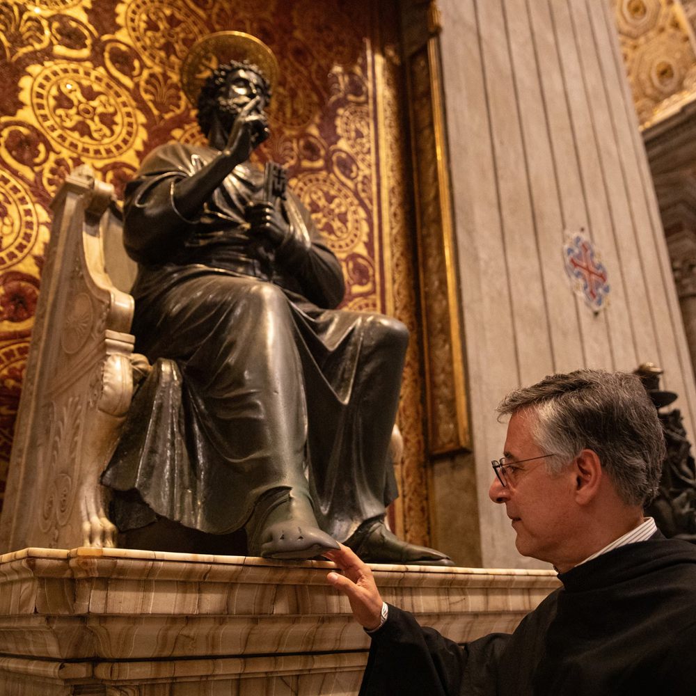 A Priest touches the foot of the famous bronze sculpture of St. Peter in the Vatican, illustrating how the bronze surface changes over centuries of human interaction.